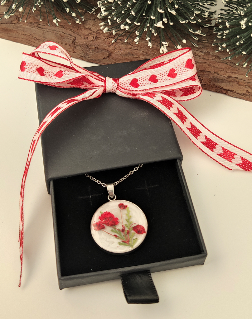 Handcrafted silver-tone necklace with a circular resin pendant featuring real pressed red flowers, presented in a black jewelry box decorated with a red and white heart-patterned ribbon