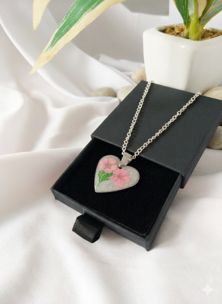 Handmade heart-shaped resin necklace with real pink dried flowers inside, placed in a luxury black gift box on a white silk background. The pendant is made of polished stainless steel.