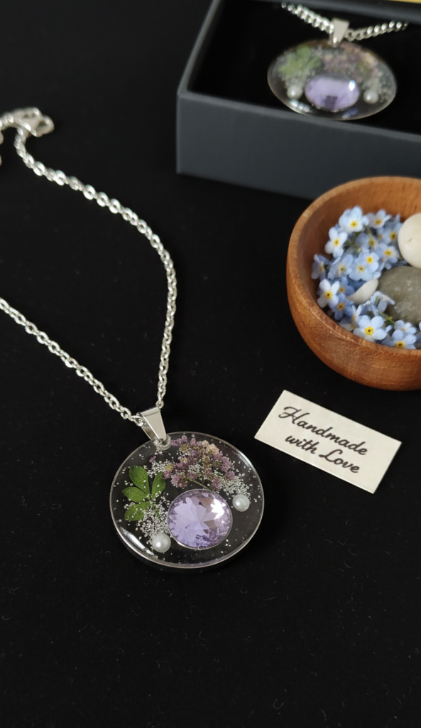 Round resin pendant with real dried flowers, tiny pearls, and a large lavender crystal. The necklace is handcrafted with stainless steel and displayed on a black surface next to a small wooden bowl of blue forget-me-nots.